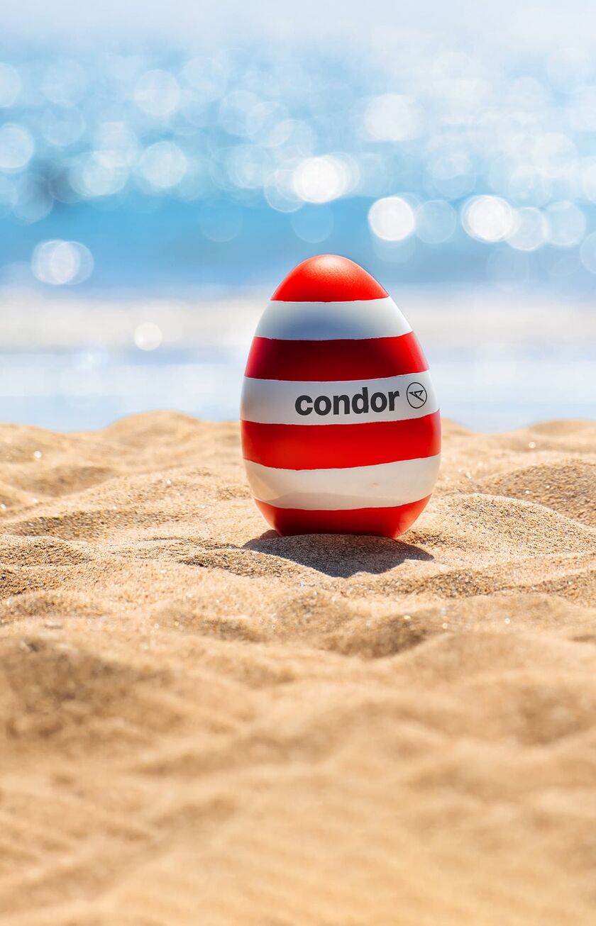 A striped Easter egg is laying in the sand, the blue sea is in the background