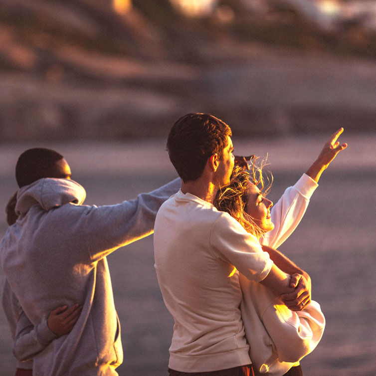 A group of friends stands on the beach at sunset, pointing at something in the distance.