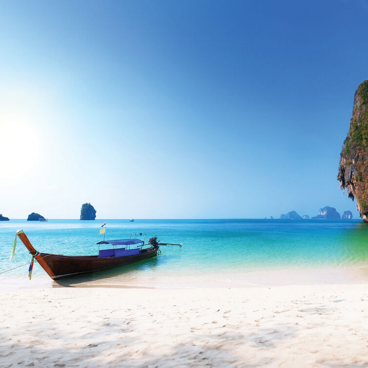  A traditional longtail boat moored on the pristine white sands of a beach in Phuket, Thailand, with crystal clear turquoise waters and limestone karsts in the distance under a bright blue sky.