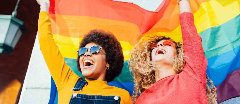 Two women in colourful clothes are holding up a rainbow flag and laugh