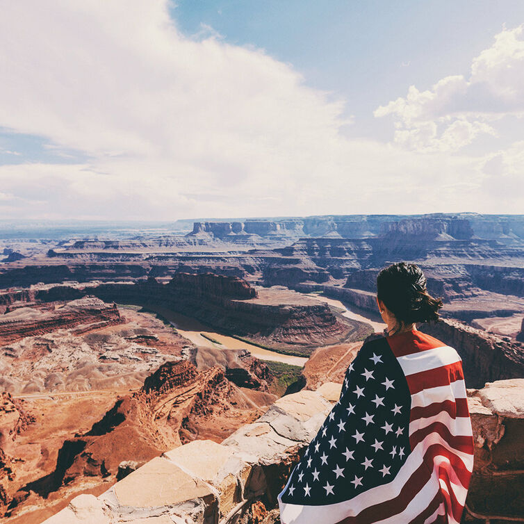 A woman is standing on top of a rock at grand canyon; she has a US flag around her shoulders.