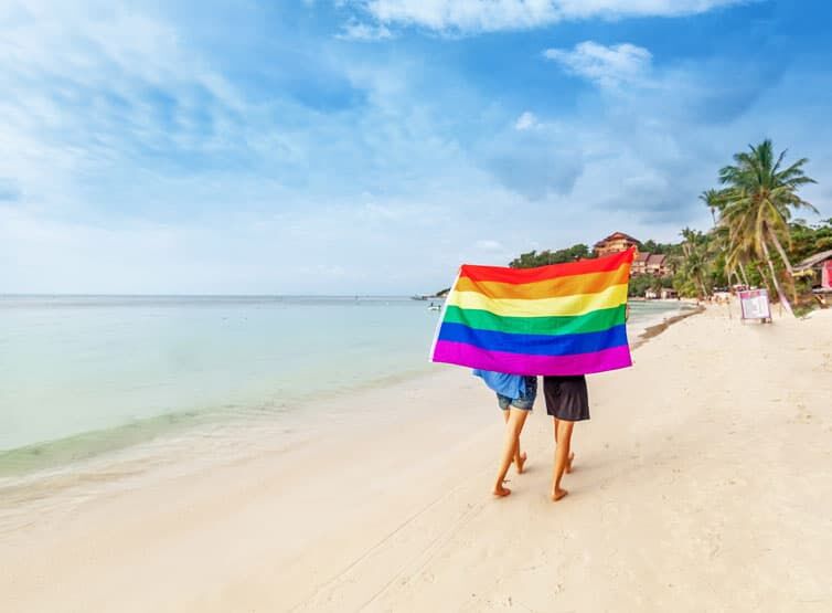 Lesbian couple walking along Caribbean beach with under Pride flag, palm trees and turquoise sea in background