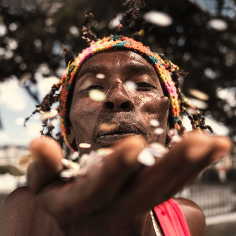 A detailed portrait of a Jamaican man blowing confetti at the camera; palmtrees are in the background