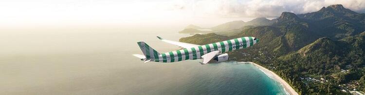 Condor airplane with a green and white striped design flying over a turquoise coastline, with lush mountains in the background during golden hour."