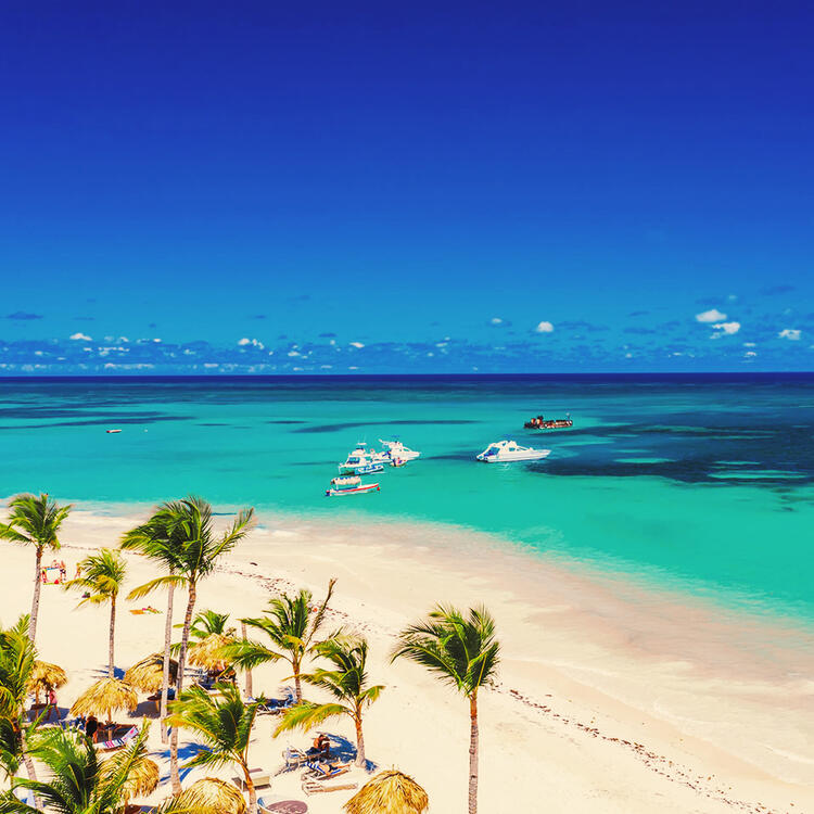 Punta Cana tropical beach with tall palm trees and thatched umbrellas lining the soft white sands, as boats float on the clear turquoise waters against a bright blue sky