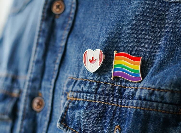 A closeup of a denim shirt with a Pride pin and a Canadian flag pin