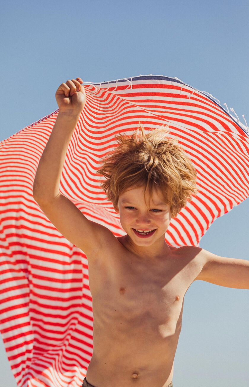 Un enfant souriant joue avec une grande serviette rayée rouge et blanche sous le soleil d’été