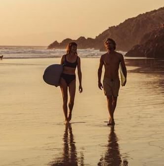 Twee surfers lopen langs het strand bij zonsondergang