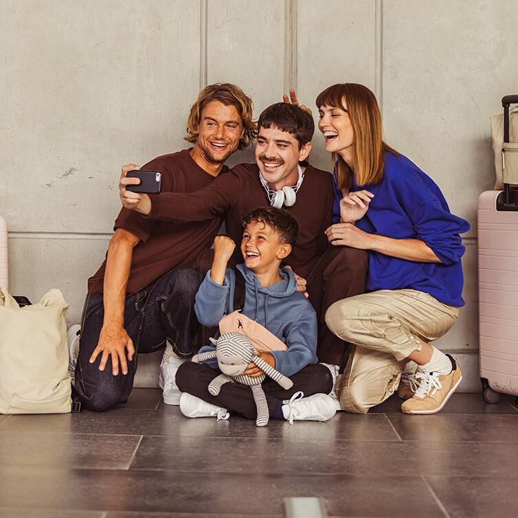 Twee mannen, een vrouw en een kind, die er gelukkig uitzien en glimlachen, nemen een selfie voor een grijze muur naast hun bagage.