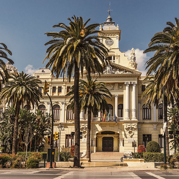 Een historisch gebouw in Malaga met klassieke architectuur, hoge zuilen en decoratieve beelden, omringd door weelderige palmbomen onder een heldere lucht.