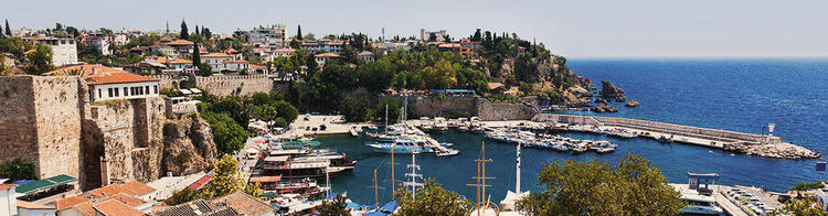  Een panoramisch uitzicht op de oude stad Antalya met historische muren, een jachthaven vol boten, en een achtergrond van groene bomen en huizen tegen een blauwe zee.