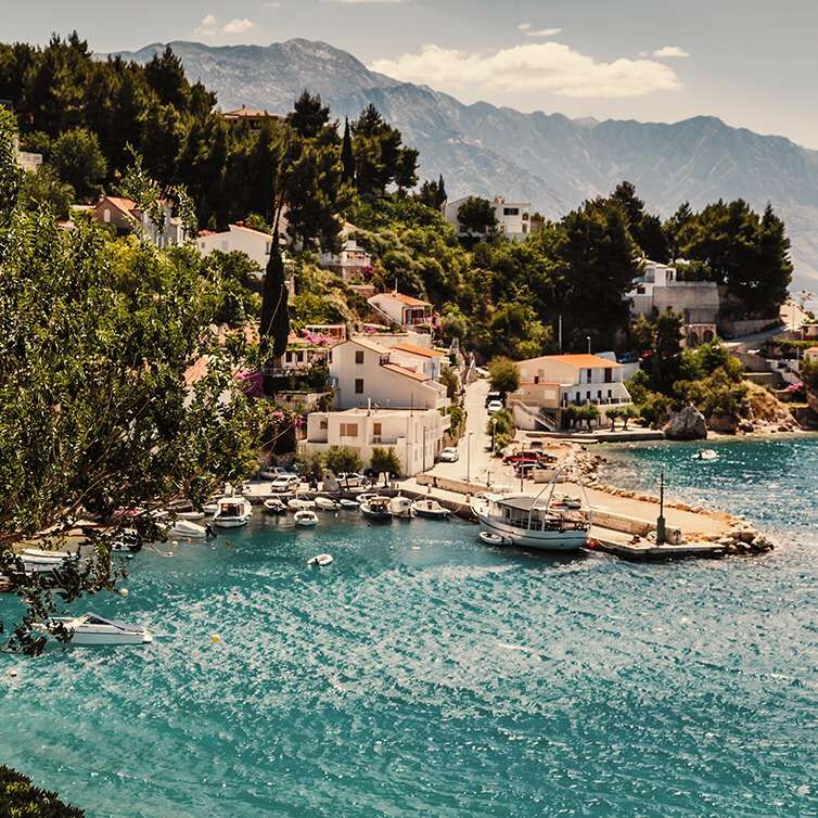 Een pittoresk kustplaatsje in de buurt van Split met pastelkleurige huizen tussen weelderig groen en uitzicht op een rustige turquoise zee met bergen op de achtergrond.