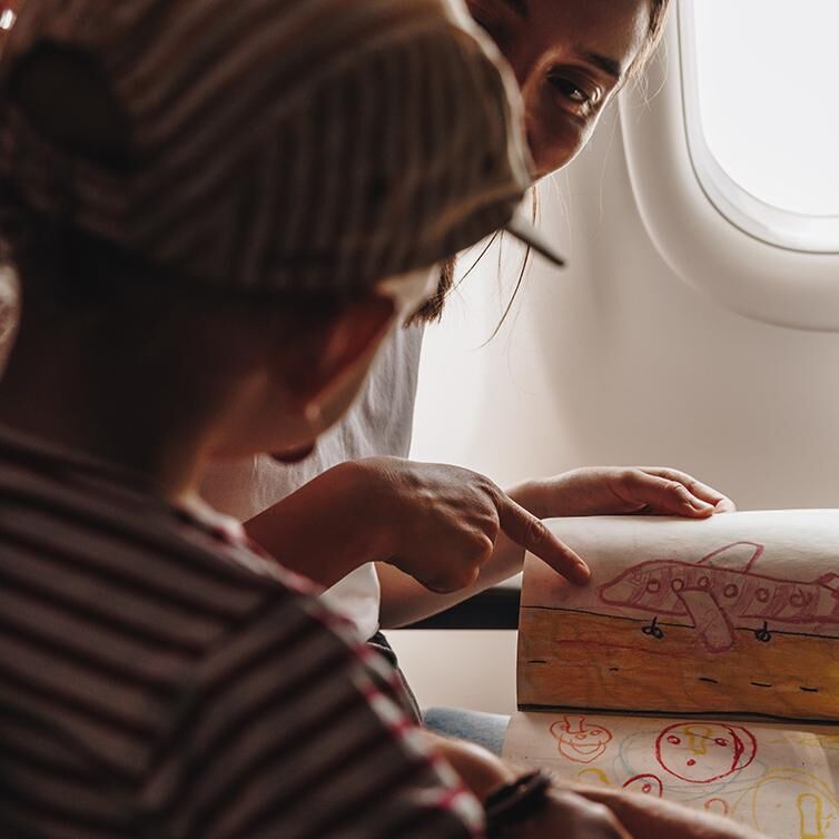 Een vrouw wijst naar kindertekeningen van een vliegtuig en emoji-achtige gezichten. De vrouw en het kind zitten naast elkaar in een vliegtuig.