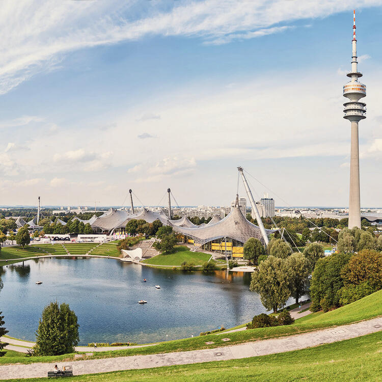 Vista aérea do Parque Olímpico de Munique, com a torre de comunicações olímpicas e a arquitetura moderna dos edifícios esportivos, cercados por áreas verdes e lagos, sob um céu claro e ensolarado.