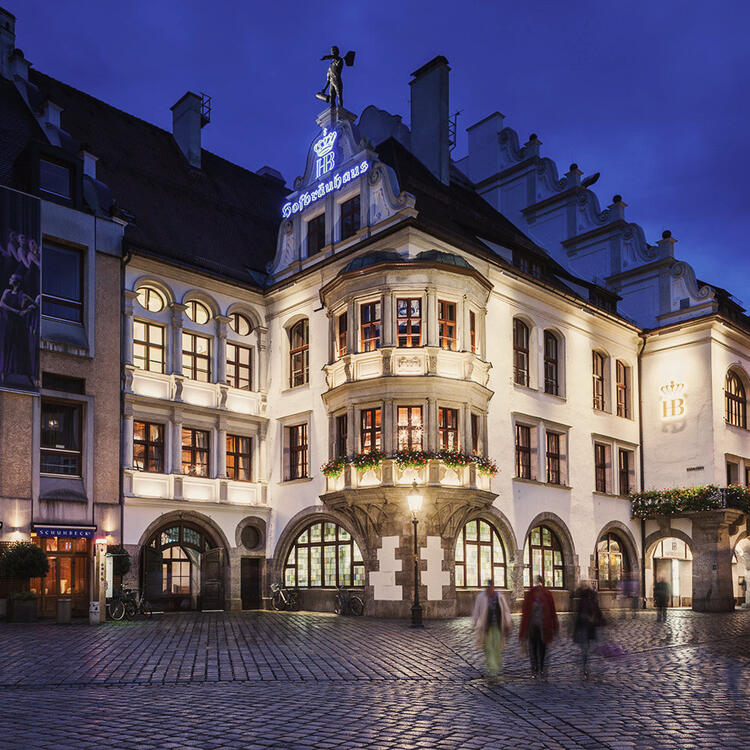 Fachada de uma cervejaria tradicional Hofbräuhaus à noite com iluminação suave e pessoas passando desfocadas em uma rua de paralelepípedos