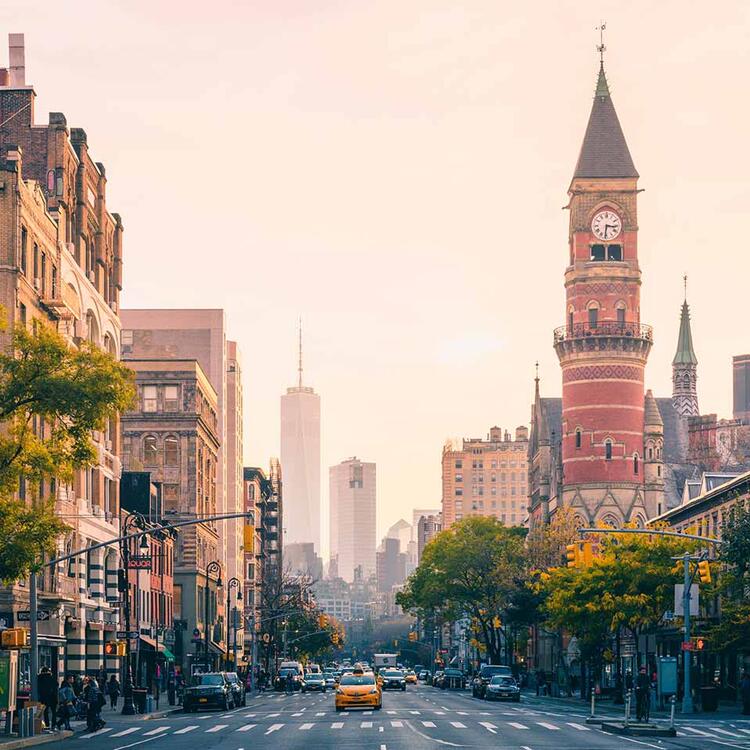 Avenida urbana em Manhattan com um táxi amarelo, prédios históricos e a torre do relógio Jefferson Market ao pôr do sol, com o One World Trade Center ao fundo.