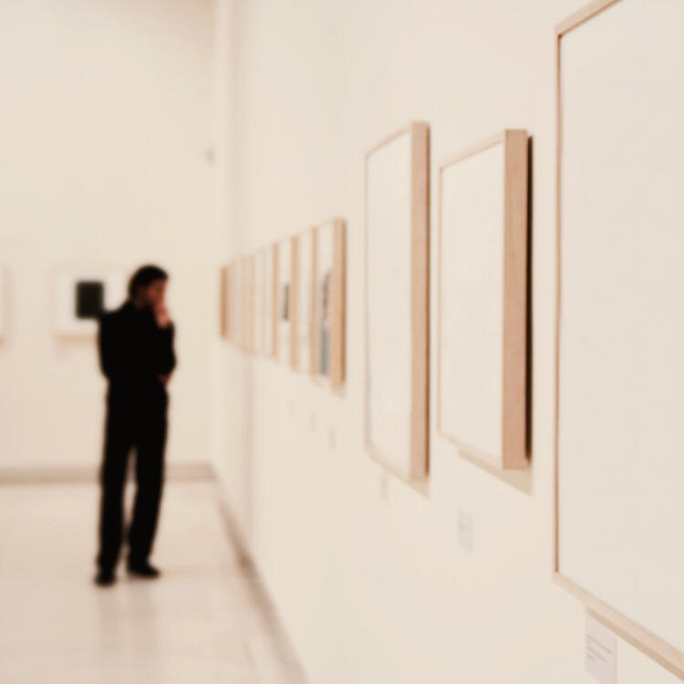 "Imagem desfocada de uma pessoa contemplando obras de arte em uma galeria de museu, com foco em quadros emoldurados alinhados na parede branca de uma sala iluminada