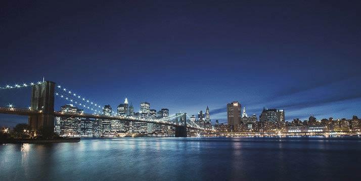 Vista noturna da Ponte do Brooklyn iluminada com o skyline de Manhattan ao fundo e reflexos das luzes na superfície da água.