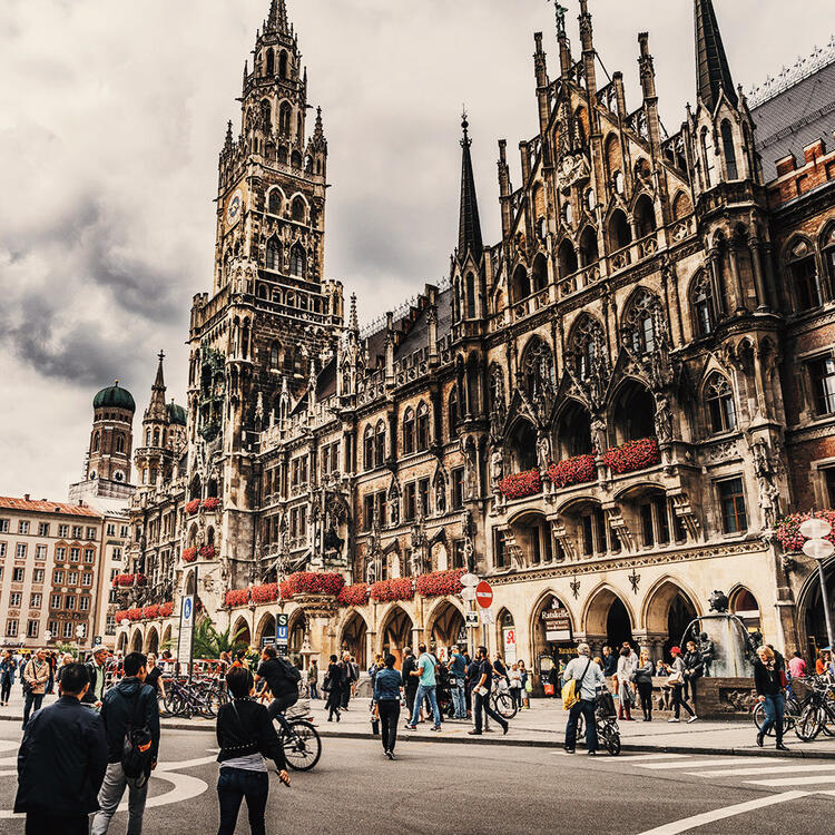 Marienplatz e a catedral em Munique | Condor Fachada gótica do Novo Câmara Municipal de Munique na Marienplatz, com pessoas e ciclistas passando em frente sob um céu nublado, demonstrando a vibrante vida urbana da praça central da cidade.