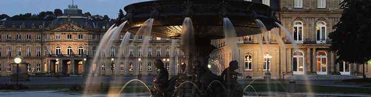 Visite Estugarda | Condor Vista panorâmica ao entardecer de uma fonte ornamental em primeiro plano, com jatos de água iluminados e esculturas detalhadas, e um edifício histórico iluminado ao fundo, sob um céu azul escurecido, sugerindo uma atmosfera tranquila em uma praça pública europeia.