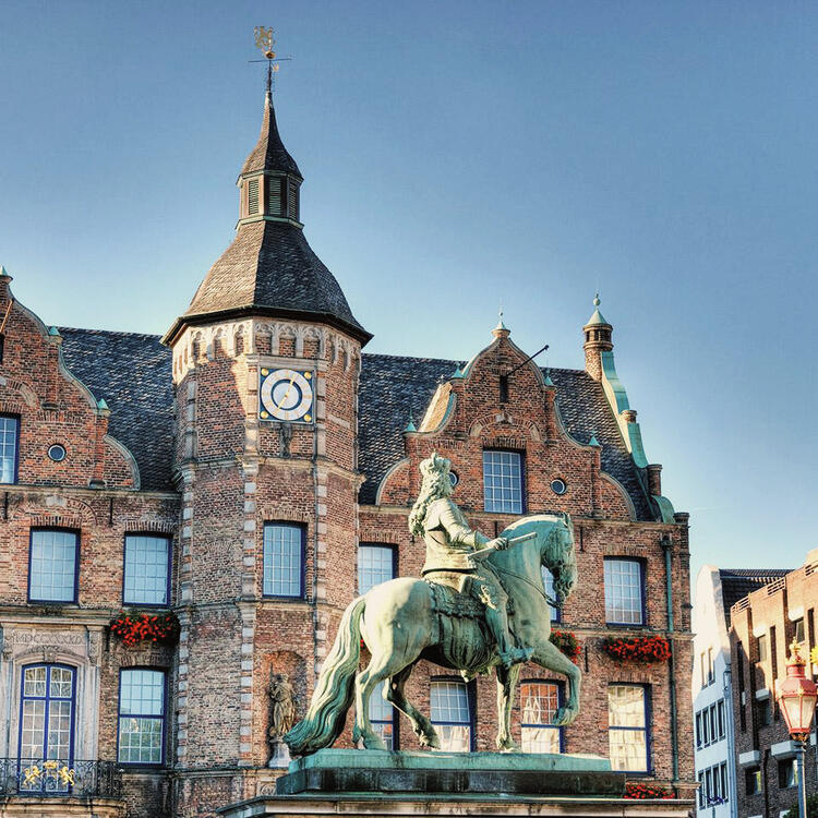 Estátua equestre em primeiro plano com um edifício histórico de tijolos ao fundo, caracterizado por um relógio de torre e decoração floral nas janelas, sob um céu azul límpido.