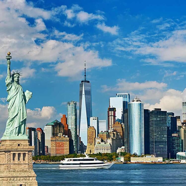 Estátua da Liberdade com a skyline de Nova Iorque ao fundo, incluindo o One World Trade Center, sob um céu azul com nuvens brancas