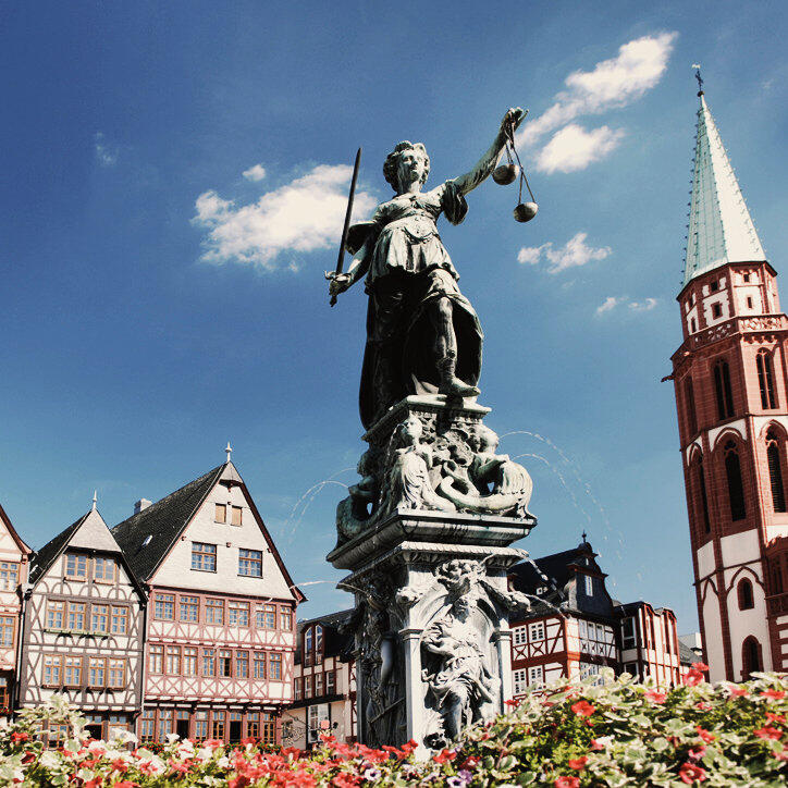 Estátua da Justiça com espada e balança em mãos, em primeiro plano contra um céu azul, com casas tradicionais em enxaimel e a torre de uma igreja ao fundo na Römerberg, a praça central de Frankfurt.