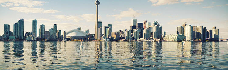 Panorama da skyline de Toronto com arranha-céus e uma torre icônica, refletida sobre as águas tranquilas, sob um céu com nuvens dispersas.