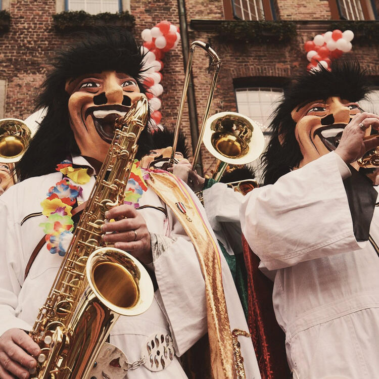 Músicos em trajes de carnaval com máscaras exageradas e coloridas tocando saxofone e trompete durante uma celebração festiva nas ruas.