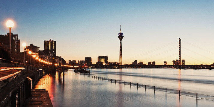 Visite o rio Reno em Düsseldorf | Condor Vista noturna do rio Reno em Düsseldorf, com a iluminação a refletir na água, a Rheinturm e a ponte suspensa destacando-se no horizonte urbano