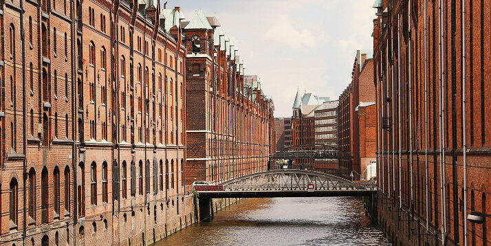 anal estreito da Speicherstadt em Hamburgo, ladeado por armazéns históricos de tijolos com ponte de metal, refletindo a herança marítima e a arquitetura industrial da cidade
