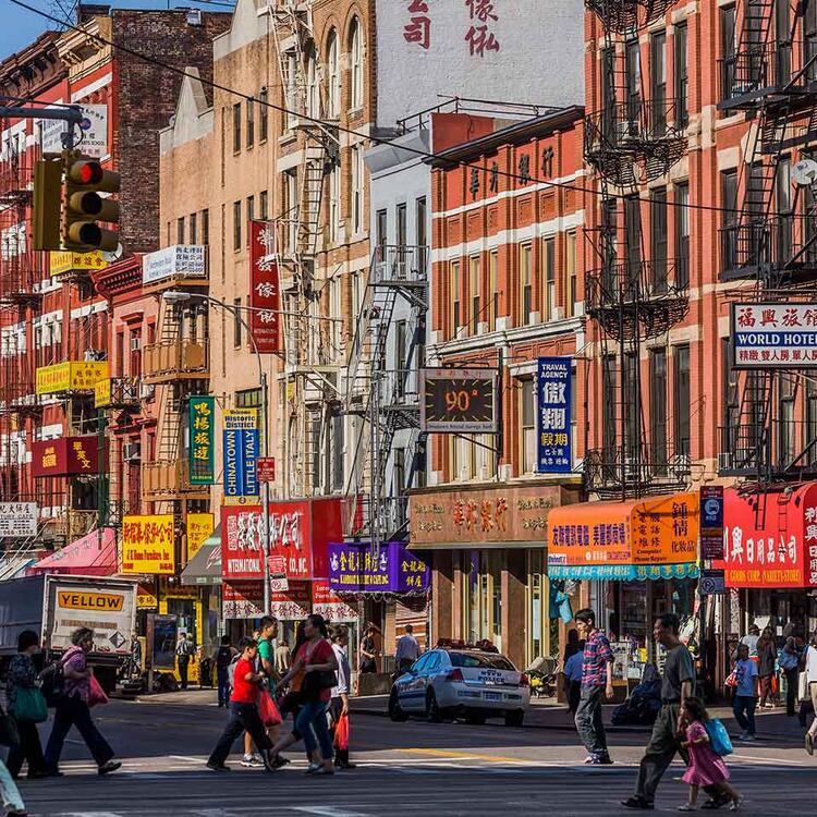 Rua movimentada em Chinatown com pedestres atravessando a rua, fachadas de edifícios com letreiros coloridos em chinês e inglês.