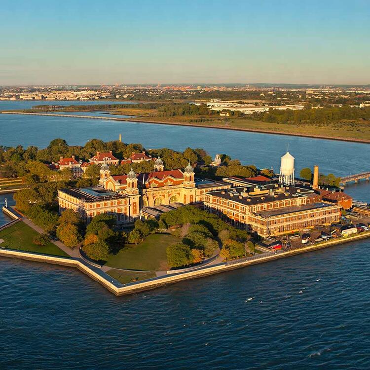 Vista aérea da Ellis Island com seu histórico edifício de tijolos e telhados de cobre ao entardecer, cercado pelas águas calmas de um rio