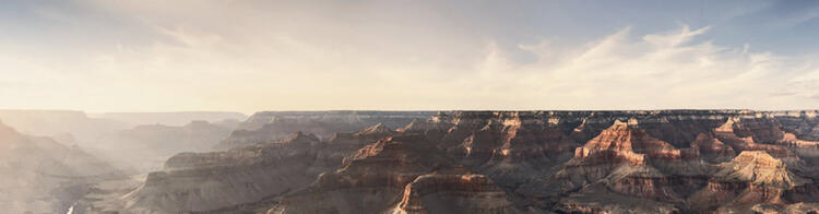 Vista panorâmica do Grand Canyon durante o pôr do sol com luz suave incidindo sobre as formações rochosas e um céu claro com nuvens delicadas