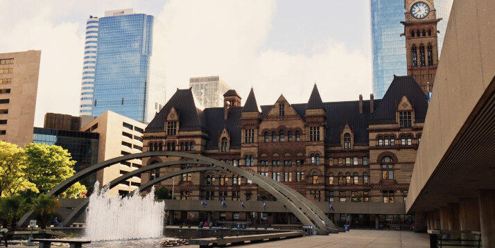 Vista da praça da prefeitura de Toronto com uma fonte e uma moderna escultura em arco, tendo ao fundo o edifício histórico do governo com uma torre do relógio, contrastando com arranha-céus contemporâneos sob um céu parcialmente nublado.