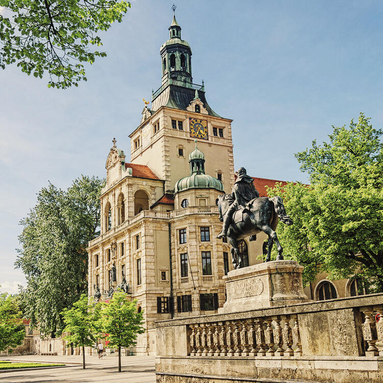 Munique e os seus museus | Condor Estátua equestre em primeiro plano com um grande edifício histórico ao fundo em um dia ensolarado, com árvores verdes e céu azul parcialmente coberto por nuvens.