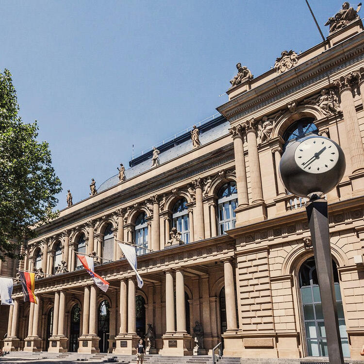 Fachada clássica da Bolsa de Valores de Frankfurt em um dia ensolarado, com colunas proeminentes e estátuas decorativas no telhado, uma grande relógio de rua em primeiro plano, e bandeiras esvoaçantes penduradas na entrada.