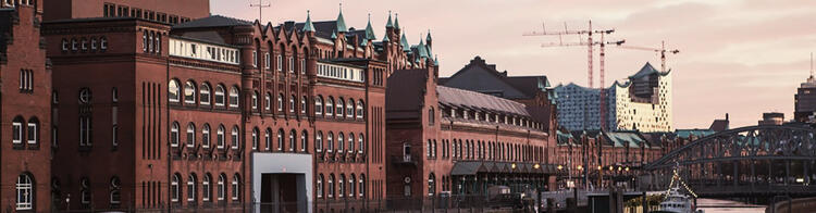 Vista panorâmica da Speicherstadt em Hamburgo ao entardecer, com seus armazéns históricos de tijolos vermelhos e telhados verdes, uma ponte de ferro e o icônico edifício Elbphilharmonie ao fundo, sob um céu rosa e azu