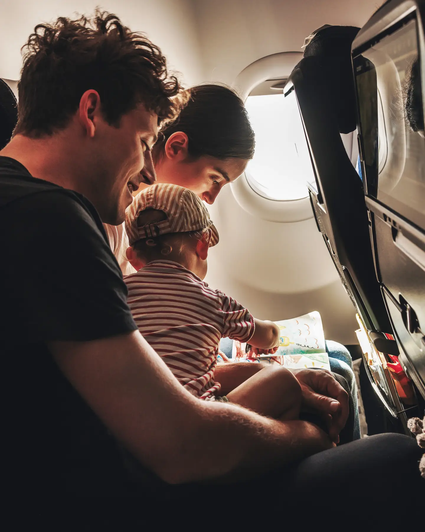 Una familia joven, formada por un hombre, una mujer y un bebé con camiseta y gorro de rayas rojas, sentados en la cabina de un Airbus A330neo. El bebé está sentado en el regazo de su padre, dibujando en un libro para colorear.