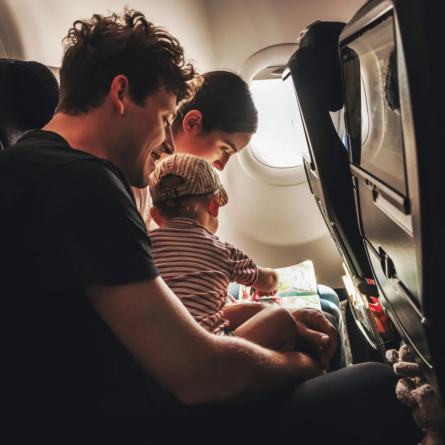 Una familia joven, formada por un hombre, una mujer y un bebé con camiseta y gorro de rayas rojas, sentados en la cabina de un Airbus A330neo. El bebé está sentado en el regazo de su padre, dibujando en un libro para colorear.