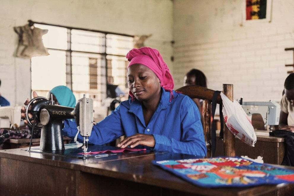Een vrouw met een roze hoofddoek zit aan een tafel en is aan het naaien