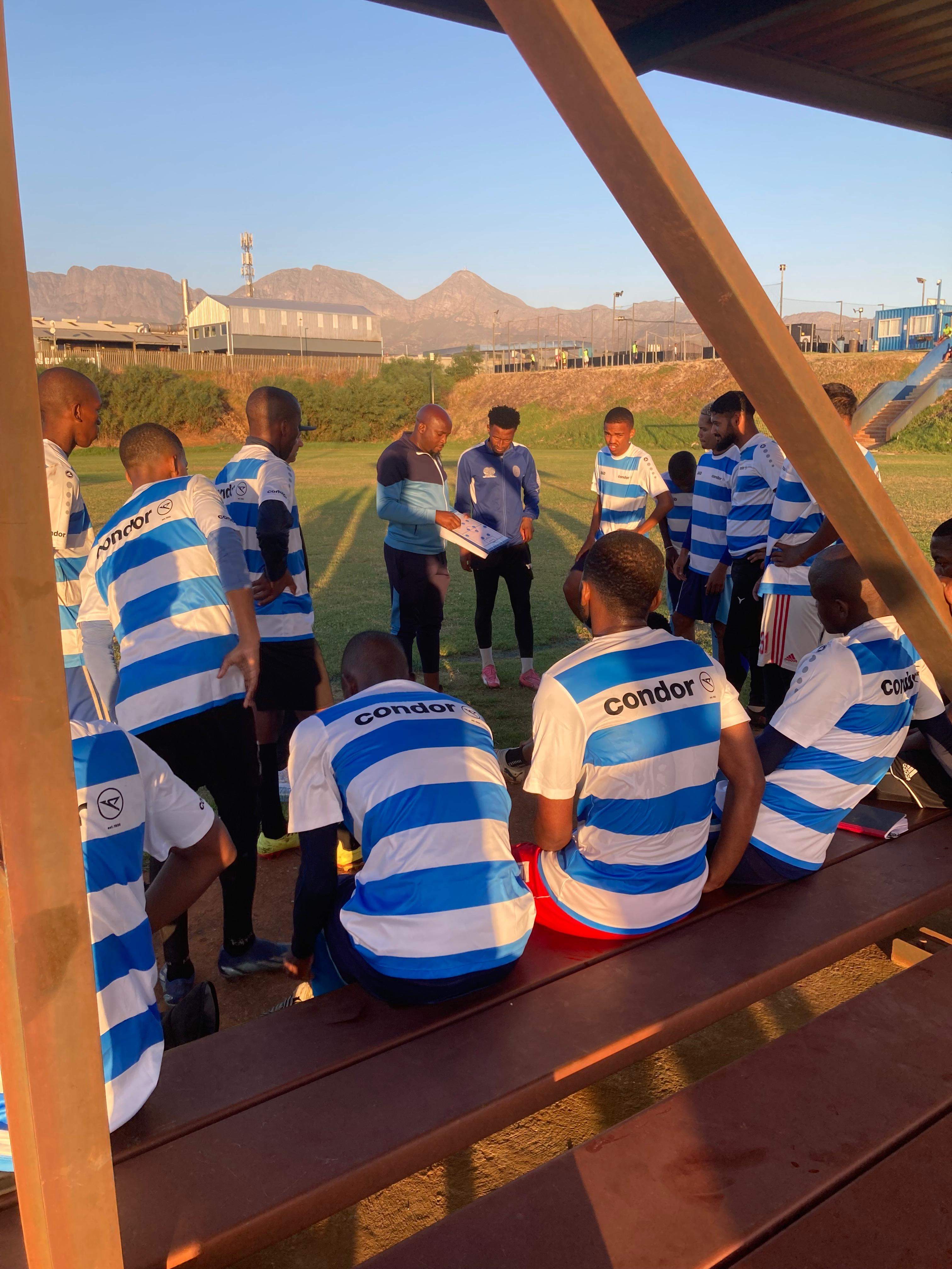 Coach briefs Young Bafana players in blue and white Condor jerseys on a field with mountain views at sunset.