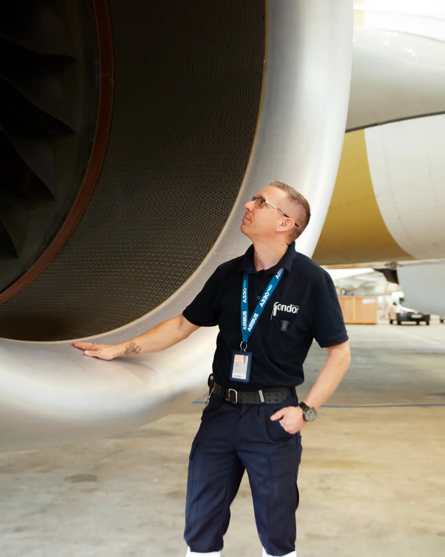 An aircraft maintenance technician in his work clothes stands in front of a large round aircraft turbine and looks inside.