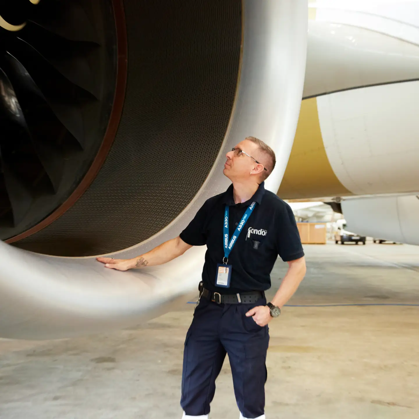 An aircraft maintenance technician in his work clothes stands in front of a large round aircraft turbine and looks inside.