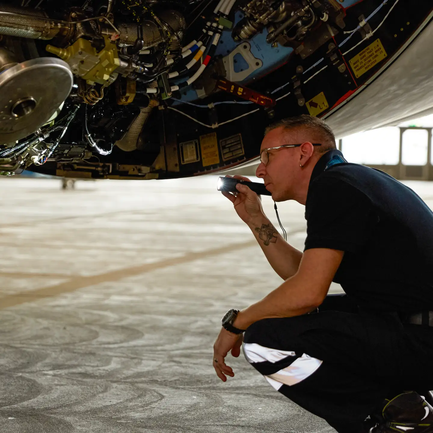 Een vliegtuigtechnicus gebruikt een zaklamp om het landingsgestel en de systeemonderdelen te controleren tijdens onderhoudswerkzaamheden in de hangar.