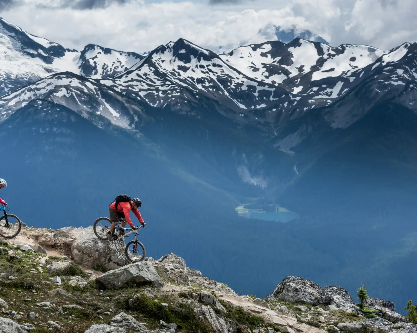 Zwei Mountainbiker auf einem Bergpfad in Whistler, Kanada, mit schneebedeckten Gipfeln im Hintergrund.