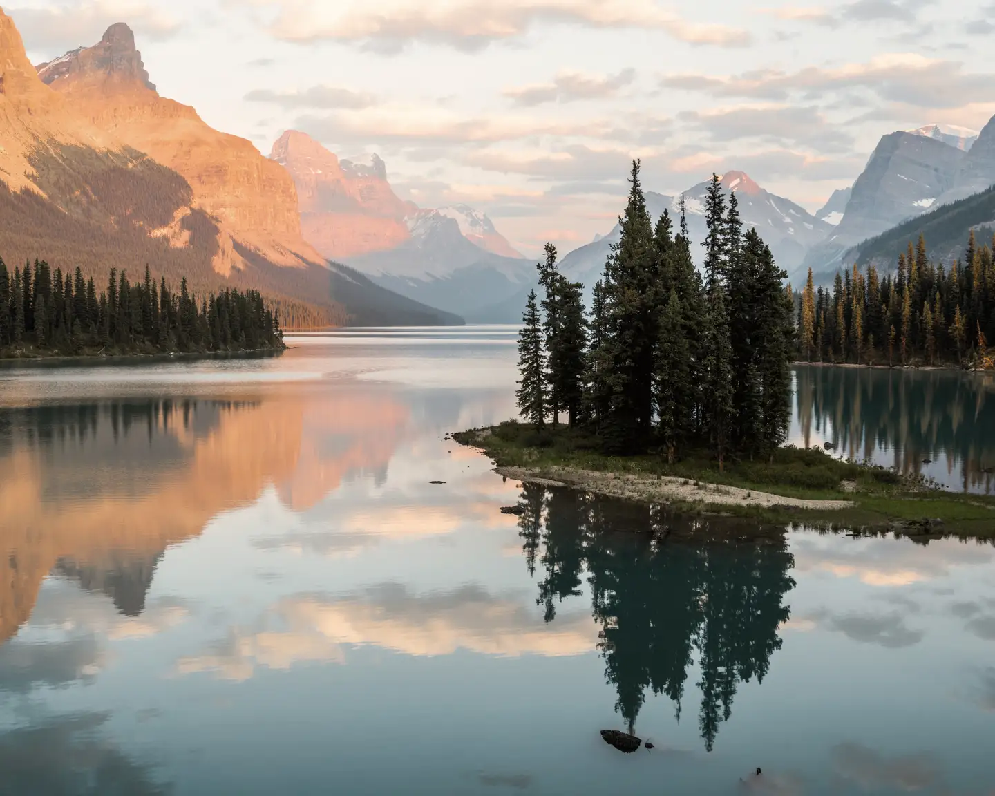 Un lac de montagne niché au cœur des Rocheuses canadiennes, avec une petite île boisée, entourée d'eaux réfléchissantes et de pics montagneux illuminés par le soleil du soir.