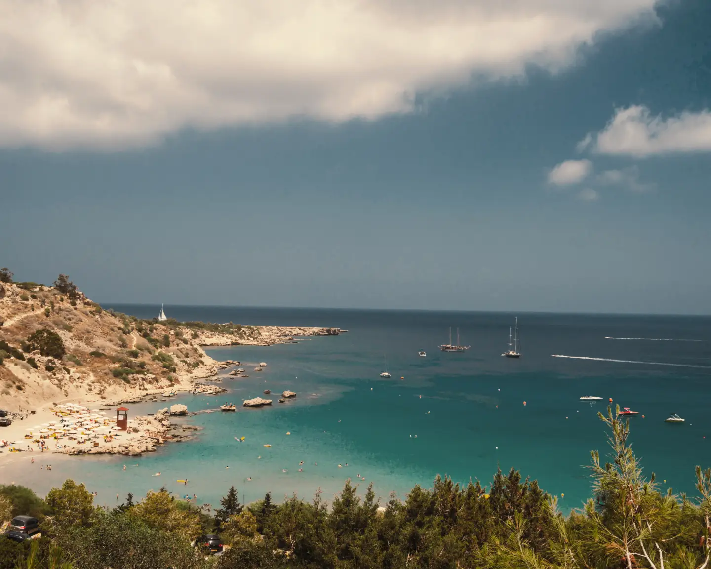 View of a beach in Larnaca, Cyprus, with clear blue water, bathers, and boats, surrounded by a hilly landscape.