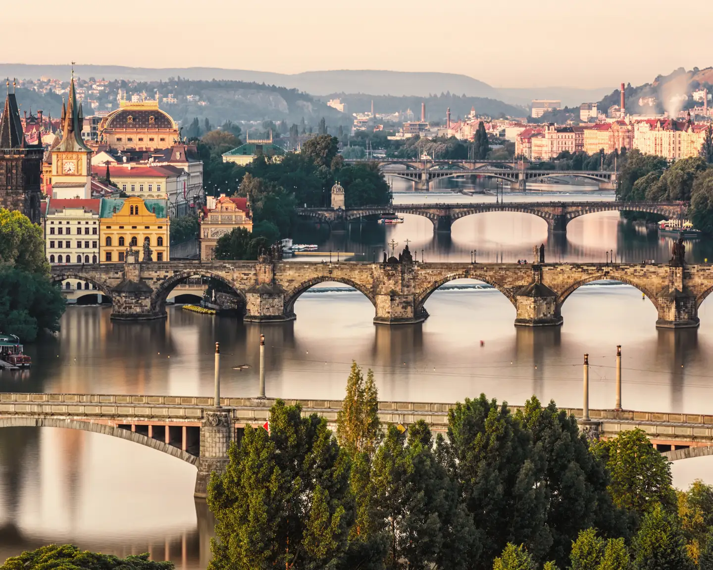 Vue du pont Charles à Prague avec la rivière Vltava et plusieurs autres ponts, entourée de bâtiments historiques et de collines verdoyantes en arrière-plan.