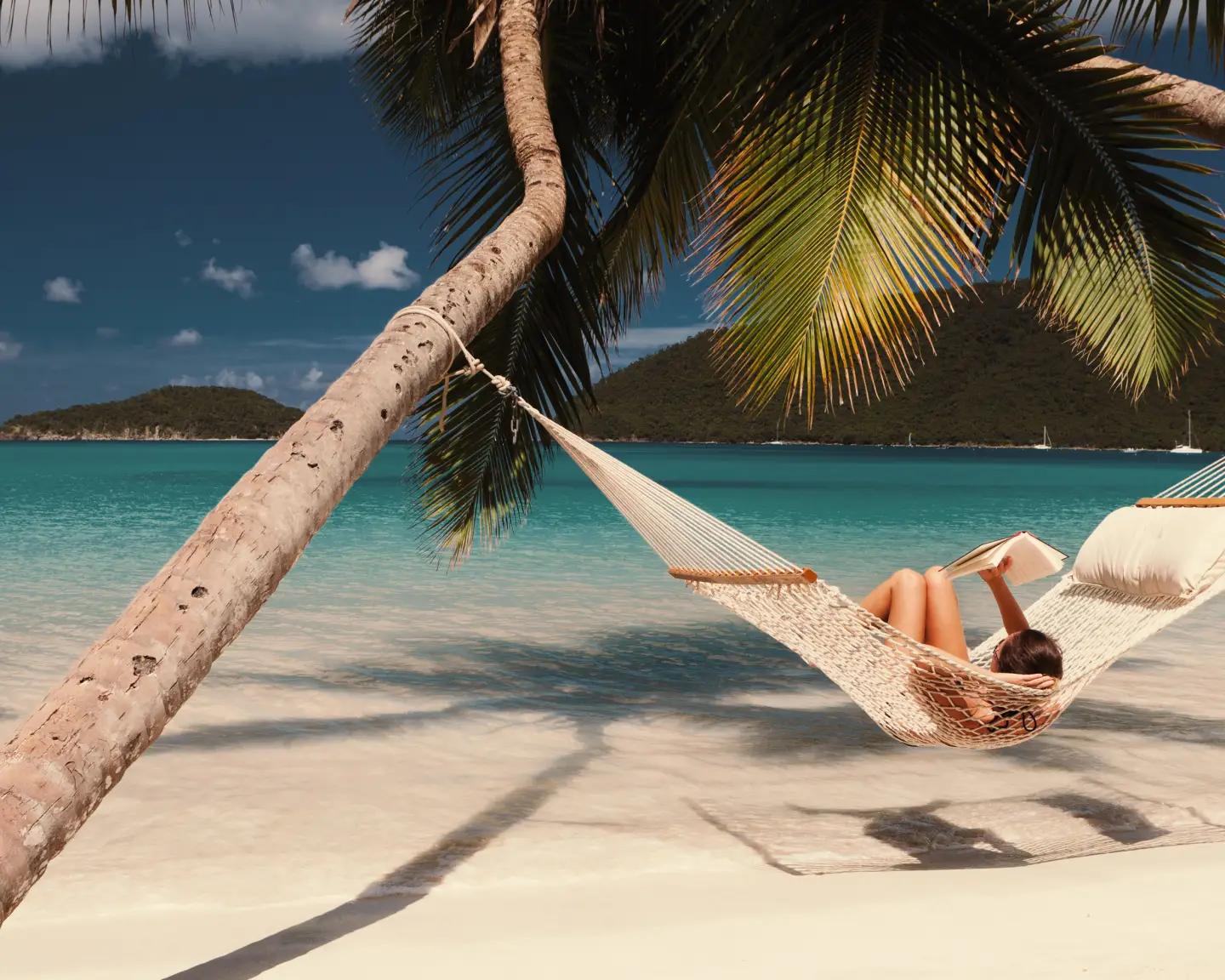 Una mujer lee un libro en una hamaca colgada entre dos palmeras en una playa tropical del Caribe.